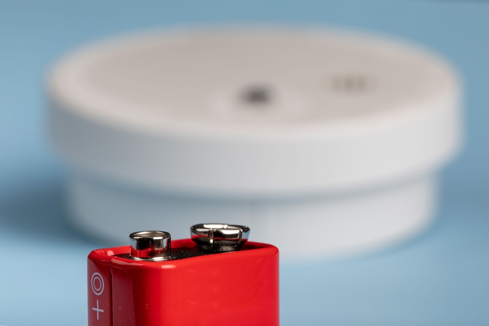Alt-text: A close-up of a red nine-volt battery in front of a blurred CO detector.